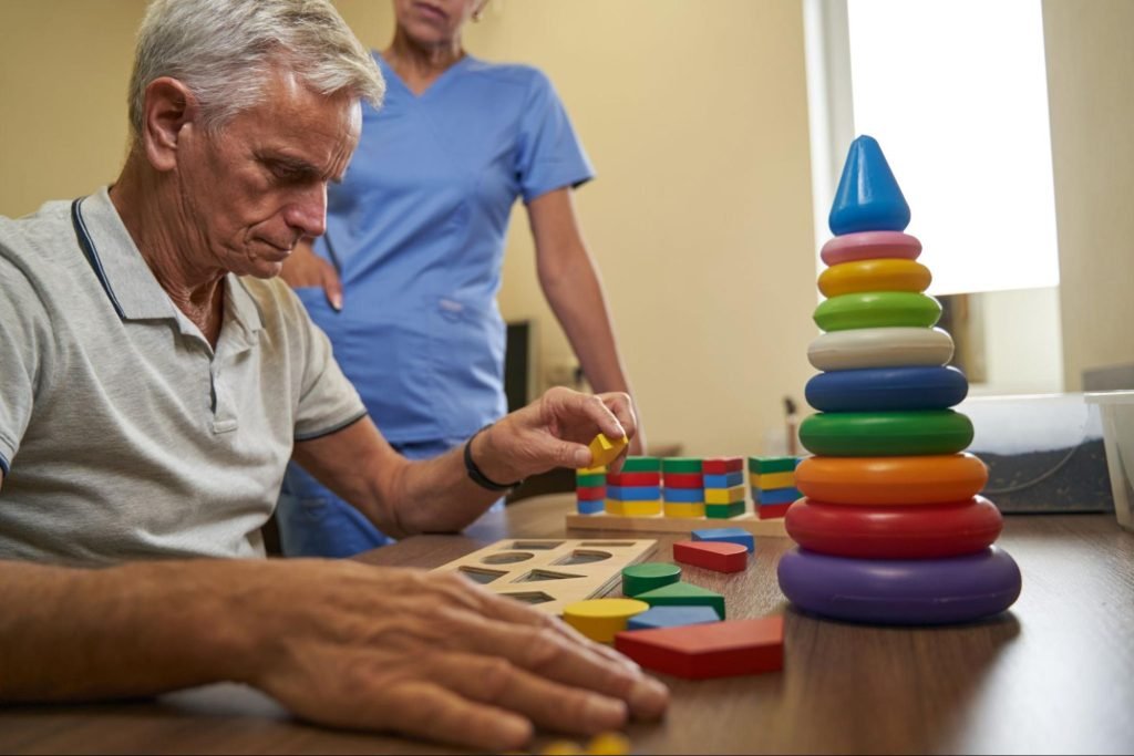An elderly patient undergoing occupational therapy plays a cognitive therapy puzzle while a nurse assists him in the background.