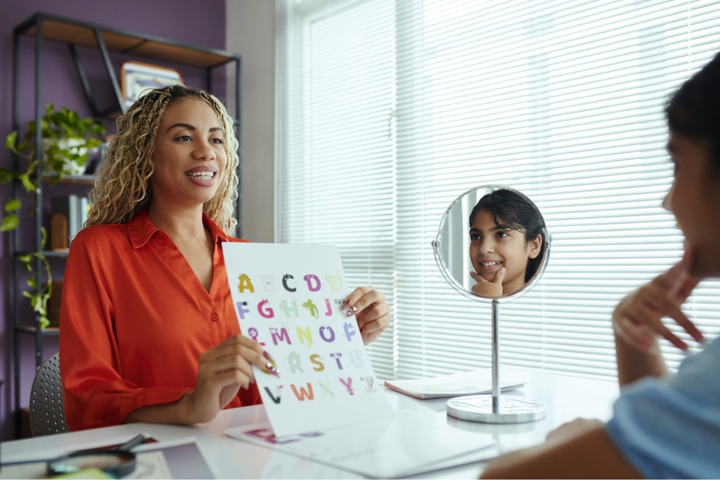 A speech therapist holds an alphabet poster while the child looks in a mirror to practice pronunciation.