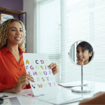 A speech therapist holds an alphabet poster while the child looks in a mirror to practice pronunciation.