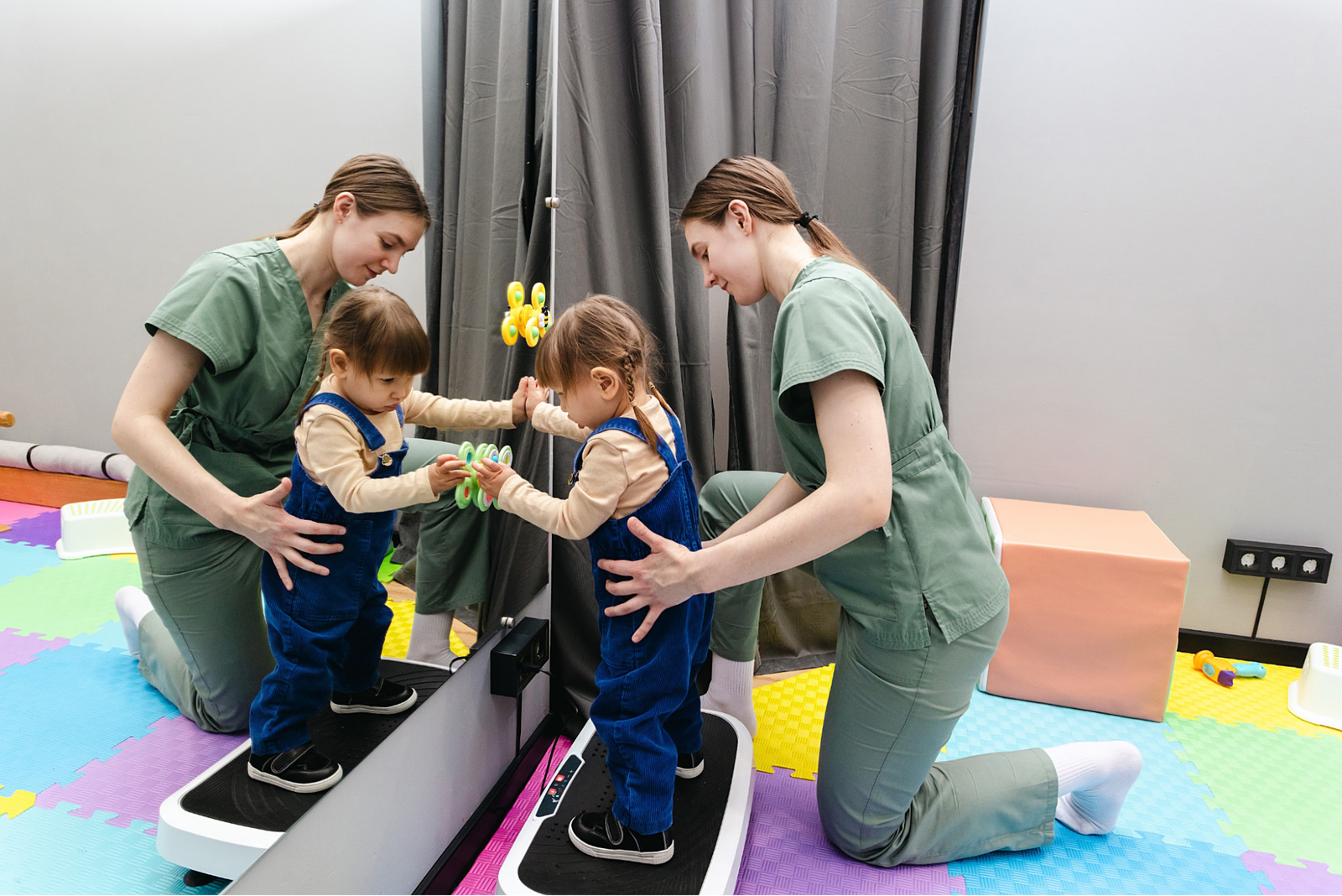 A physiotherapist assists a toddler step onto a balance training machine in front of a mirror.