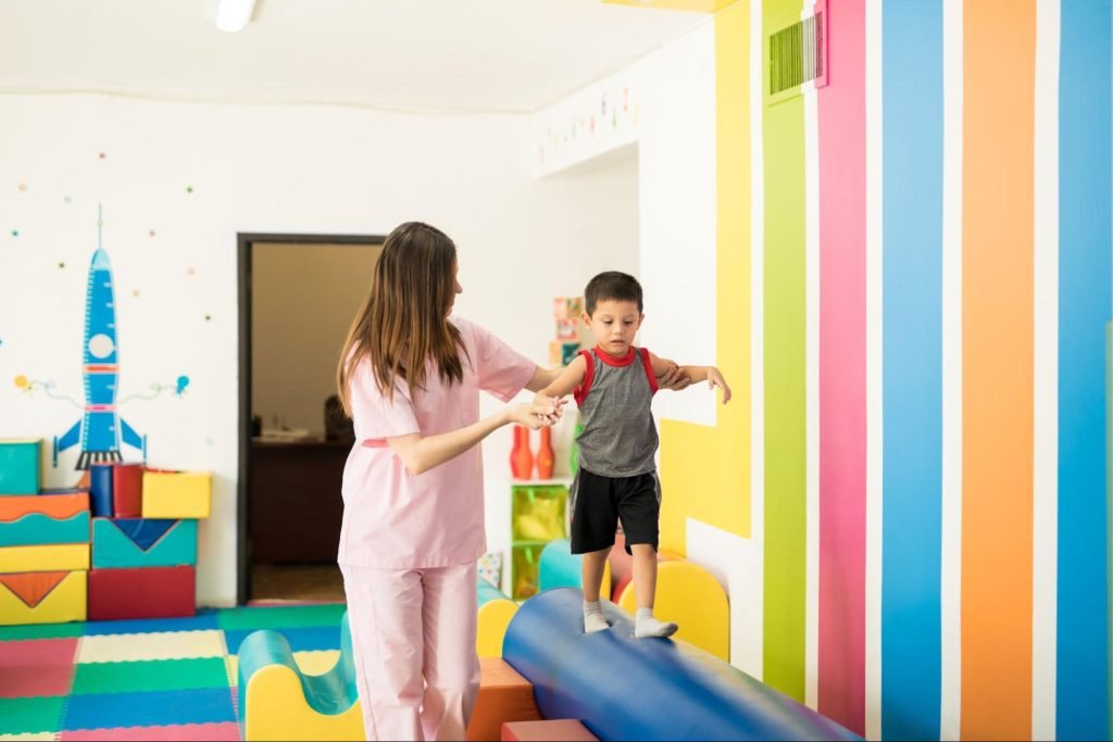 An occupational therapist guides a little boy on a foam beam for balance training.