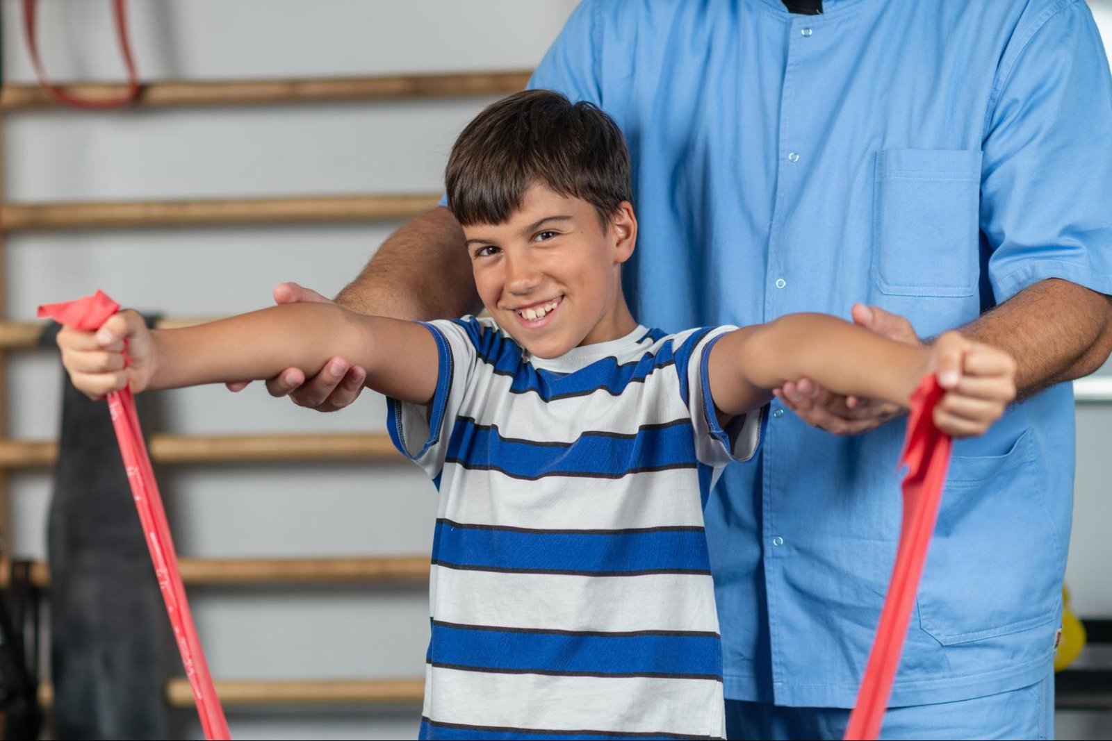 A boy undergoing physical therapy smiles at the camera while exercising his arms with an elastic band.