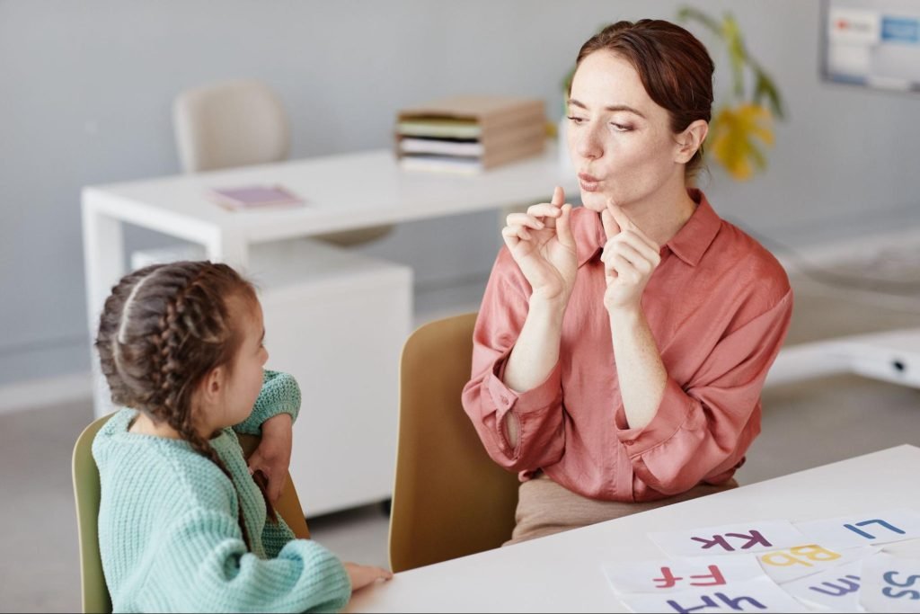 A speech therapist is sitting at a table with a toddler and is practicing mouth exercises.